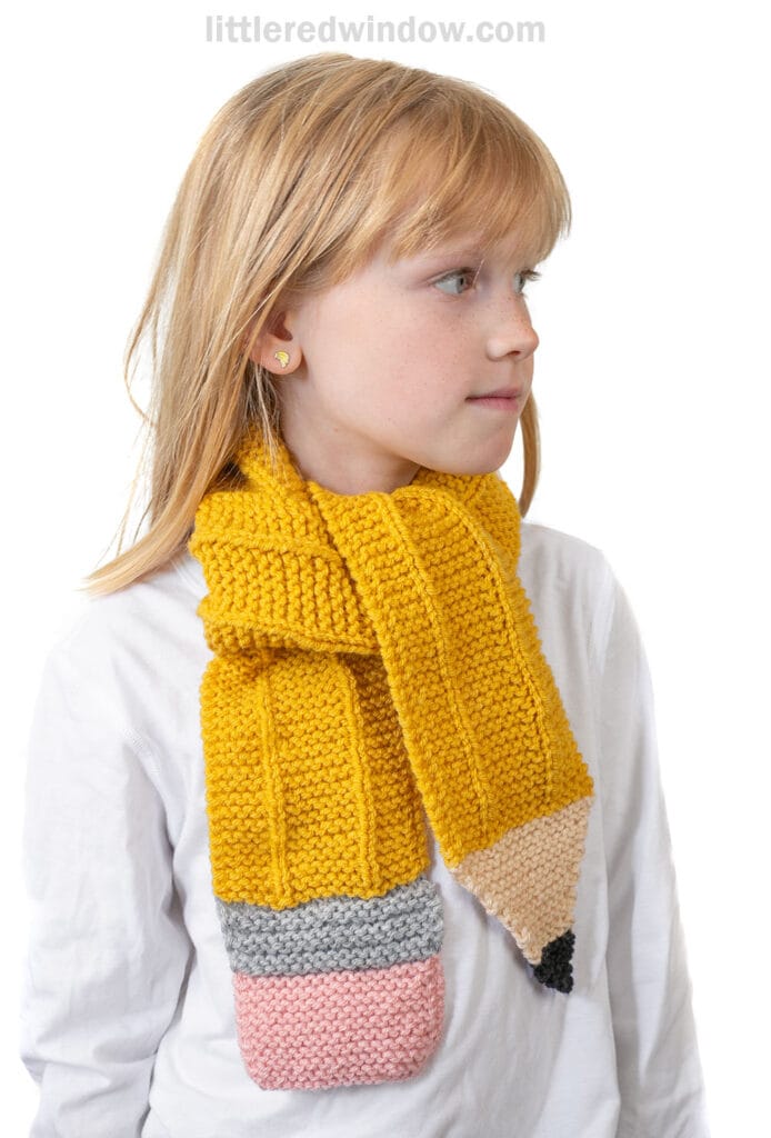 A young girl wearing a white shirt and a knitted scarf, designed to look like a yellow pencil with a pink eraser end and pointed tip, stands facing sideways against a white background.