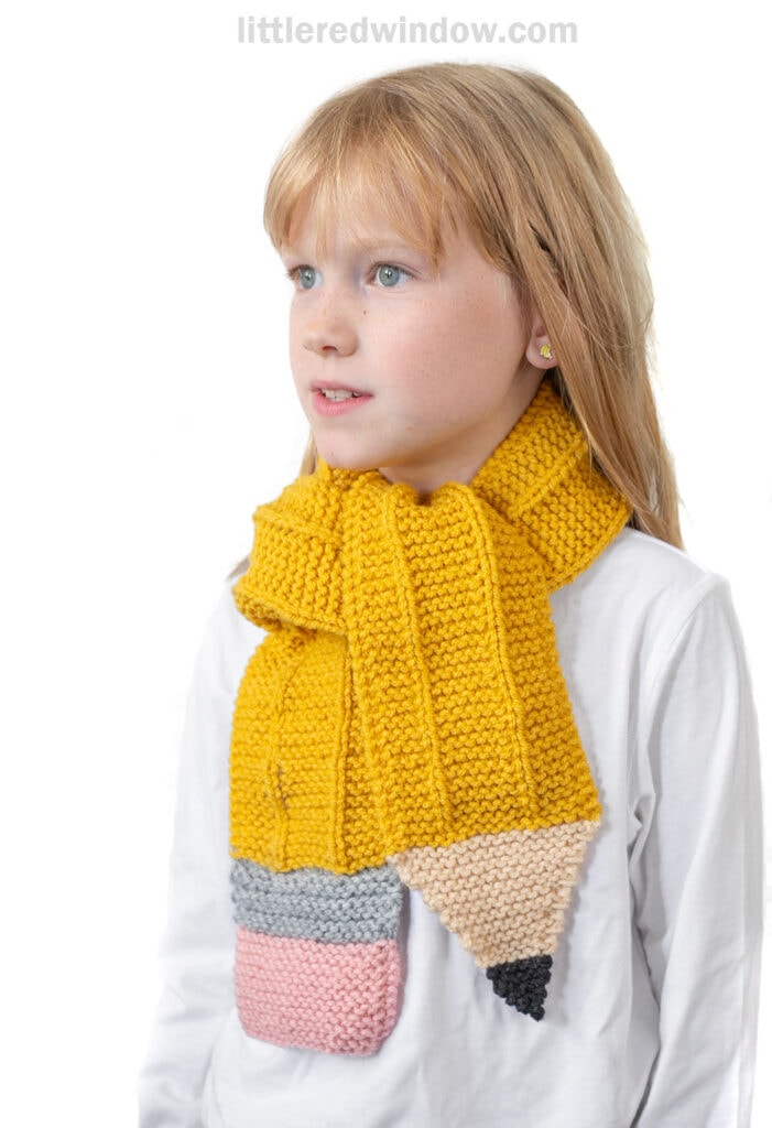 A young child with blonde hair wears a knitted yellow pencil scarf, complete with a pink eraser, gray band, yellow body, and beige tip with black point. The child is facing slightly to the right.