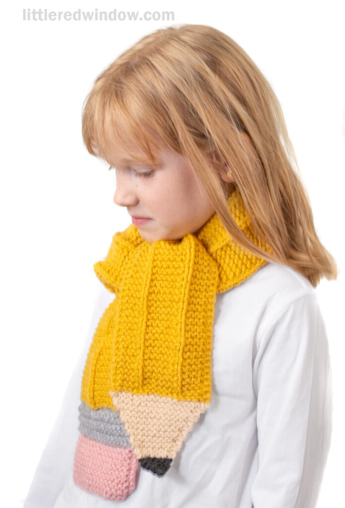 A young girl with blonde hair wears a white shirt and a knitted scarf designed to look like a yellow pencil, complete with gray, pink, and black ends. She is looking down against a white background.