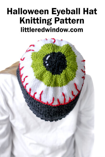 Person wearing an eyeball hat—a knitted hat designed to look like a green and black eyeball with red veins—viewed from above. Text above reads: “Halloween Eyeball Hat Knitting Pattern littleredwindow.com”.