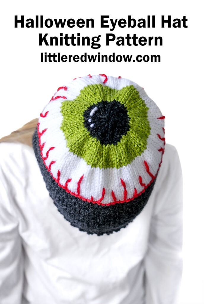 Person wearing an eyeball hat—a knitted hat designed to look like a green and black eyeball with red veins—viewed from above. Text above reads: “Halloween Eyeball Hat Knitting Pattern littleredwindow.com”.