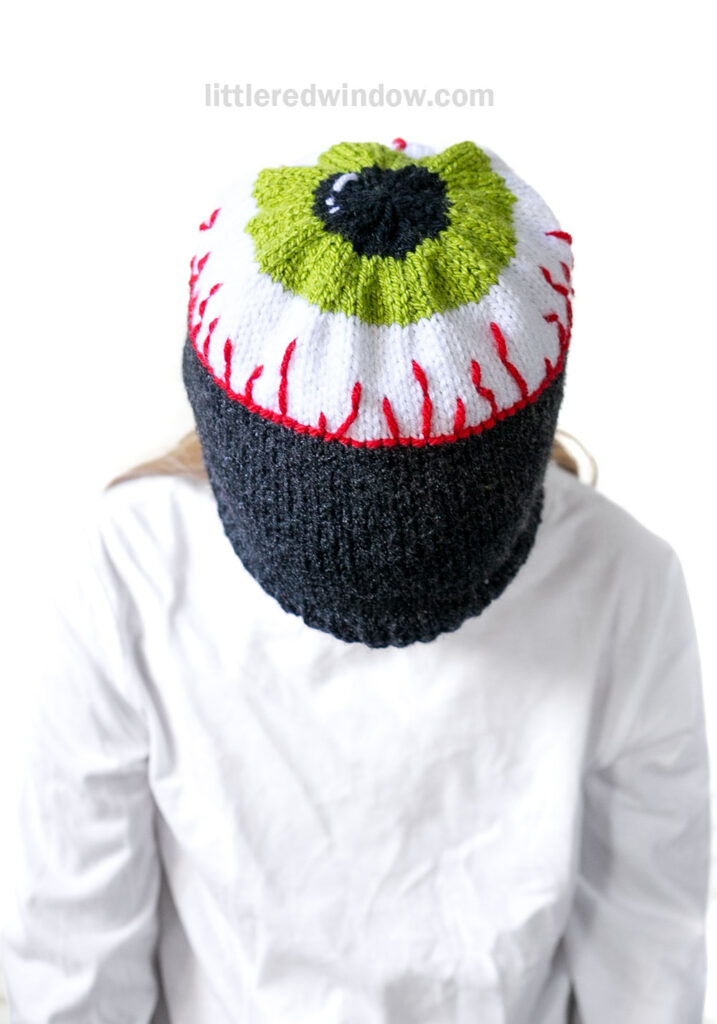Person wearing an eyeball hat—a knit hat designed to look like a large green and white eyeball with red veins—covering their entire head and face.