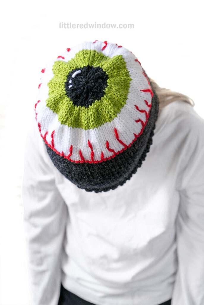 A person wearing an eyeball hat—a knit hat designed to look like a green and black eyeball with red veins—is viewed from above.