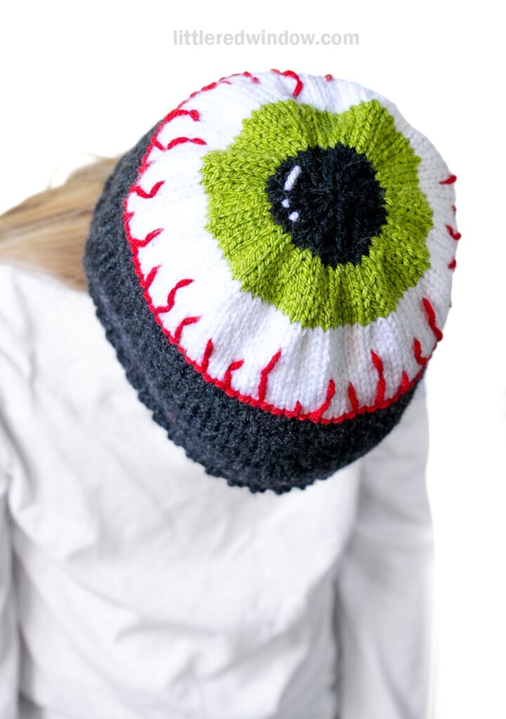 A person wearing an eyeball hat designed to look like a green eyeball with red veins is facing downward, with the quirky knit hat covering the top of their head.
