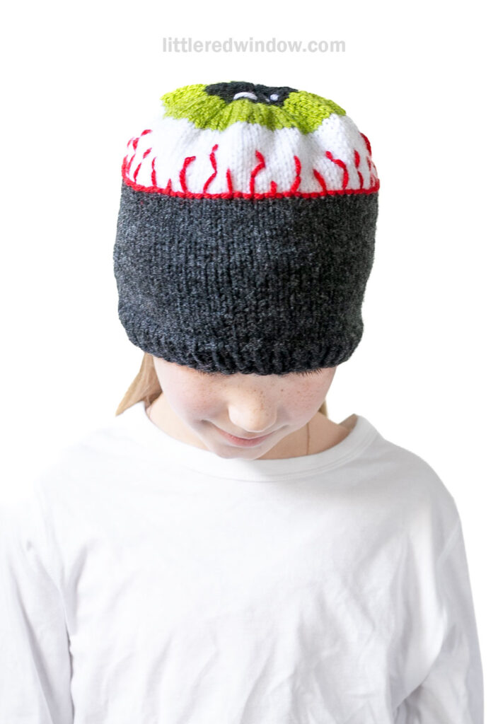 A child wearing a white shirt has an eyeball hat pulled low, covering their eyes. The knit hat is designed to look like a green and red bloodshot eyeball.