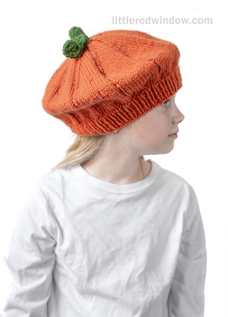 A child wearing a white shirt looks to the side, modeling an orange knitted pumpkin beret shaped like a pumpkin with a small green stem on top.
