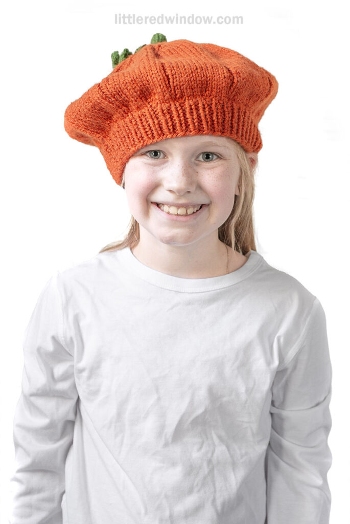 Smiling child wearing a white shirt and a playful pumpkin beret, standing against a plain white background.