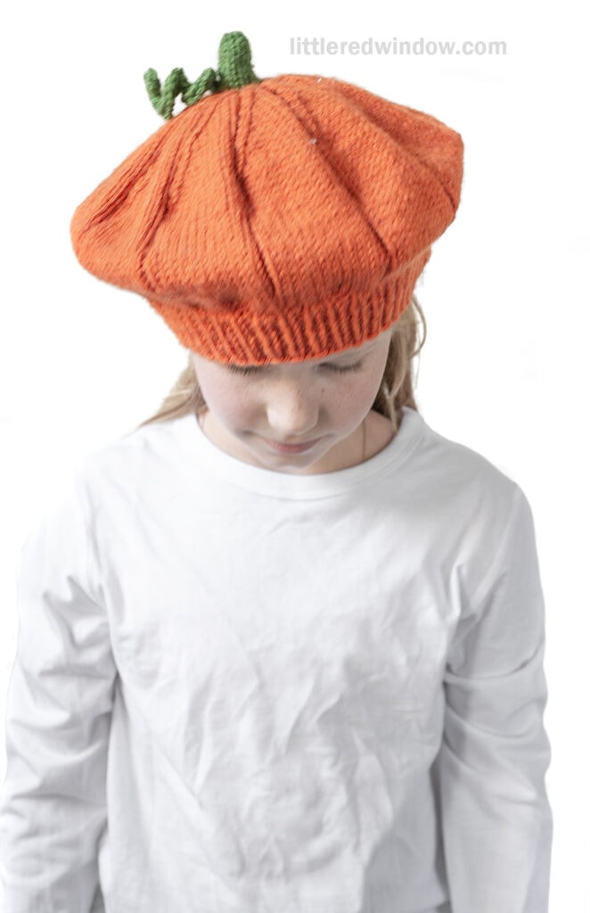 A child wearing an orange pumpkin beret, complete with a green stem on top, looks down while dressed in a plain white long-sleeve shirt.