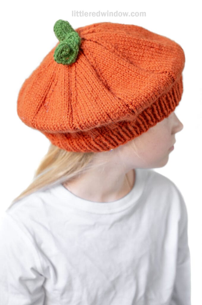 A person wearing a knit orange pumpkin beret designed to look like a pumpkin, complete with a small green stem on top, and dressed in a plain white shirt.